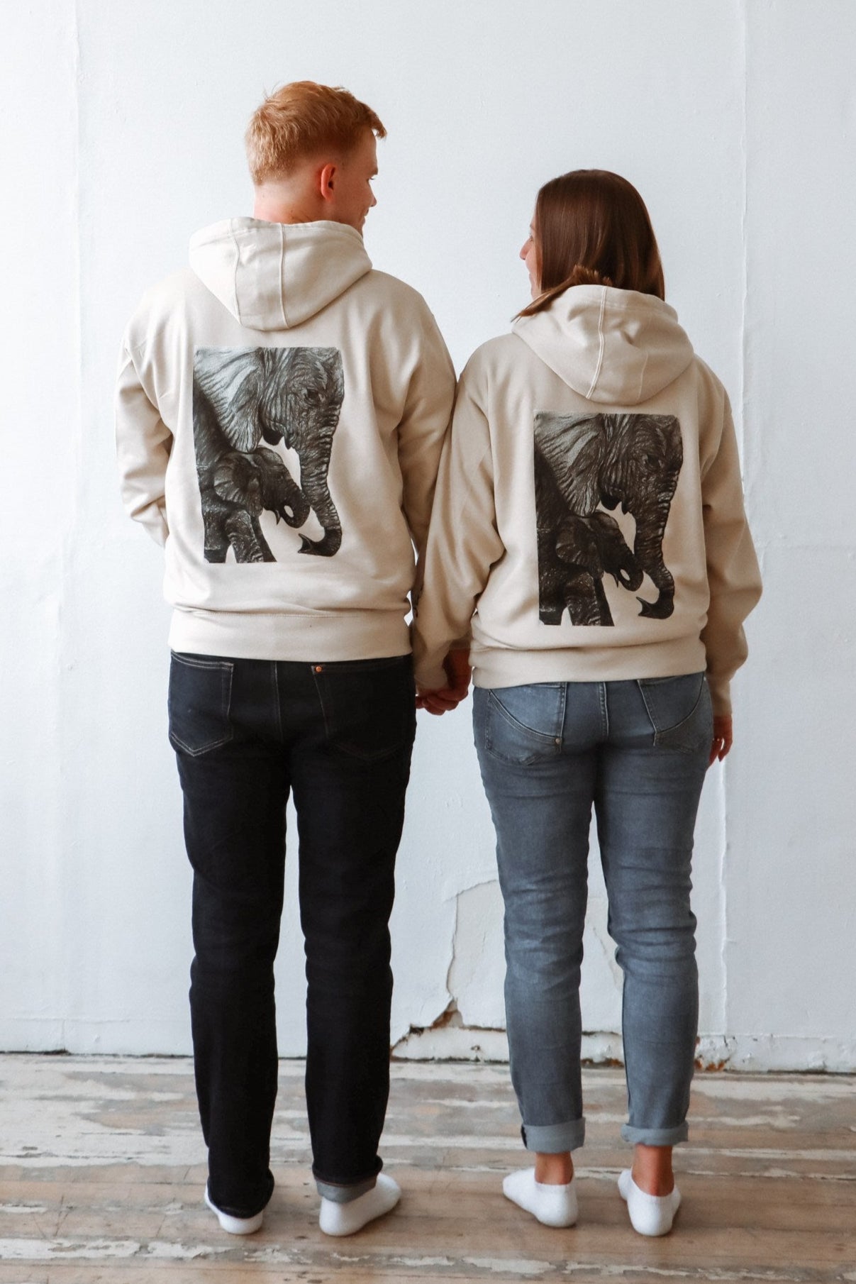 A couple holding their hands and looking at each other with love while wearing 2 beuatiifuly designed niiberg hoodies with elephants sketches on their backs of the hoodie.